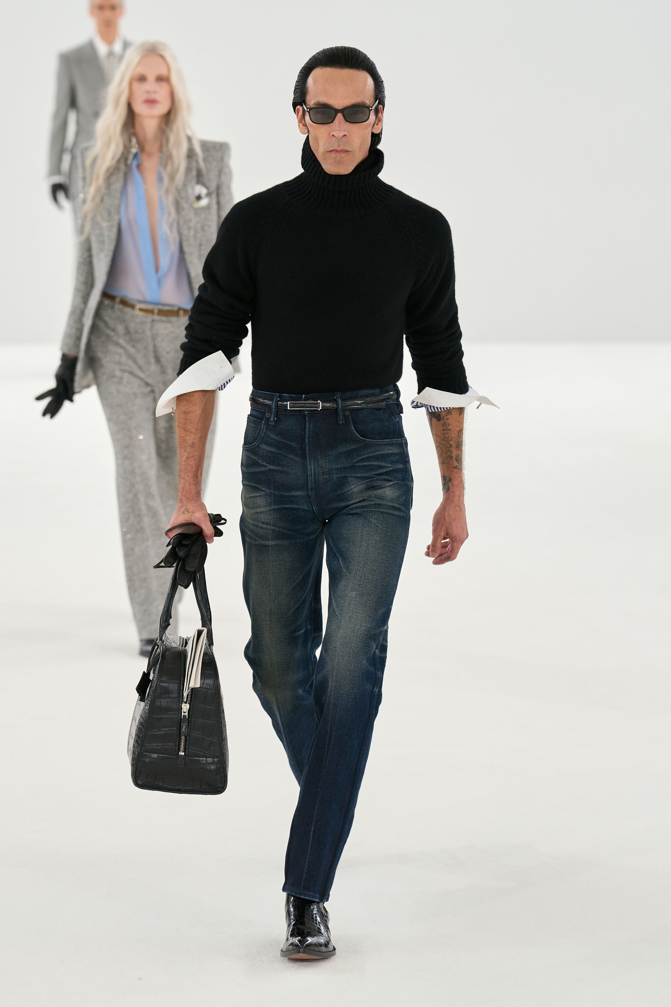 Tom Ford 2026秋冬系列回归品牌标志性的性感极简主义。高腰铅笔裙、透明材质层叠与利落西装展现强烈的都市魅力。黑色、棕色与金属光泽构成成熟而冷峻的色彩体系，皮革与光泽面料的运用强化性感张力。整体造型强调身体线条与权力感，展现Tom Ford一贯的夜晚都市美学-顽石频道趋势研究